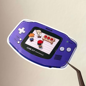 Può includere: Un adesivo viola Game Boy Advance con una scena a cartoni animati di tre personaggi e un tavolo con tre palline rosse. L'adesivo presenta il testo "GAME BOY ADVANCE" e il logo Nintendo.