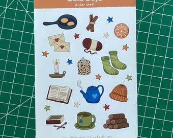 Op de afbeelding: Een stickerblad getiteld "Cold Days" met illustraties van gezellige items. Bevat een koekenpan met eieren, kaneelstokjes, sterren, een sinaasappelschijfje, enveloppen, garen, sokken, een kaars, een boek, een theepot, een muts, een theekopje en houtblokken.