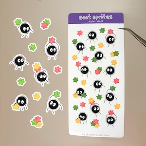 Könnte beinhalten: Ein rechteckiger Aufkleberbogen mit der Aufschrift soot sprites sticker sheet, einem violetten Kopfbereich und weißem Hintergrund. Er zeigt mehrere schwarze, kreisförmige Rußmännchen mit großen weißen Augen, die kleine rosa, grüne und gelbe Sterne halten.