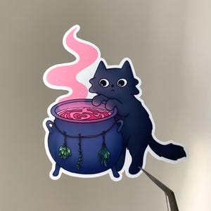 Könnte beinhalten: Ein Aufkleber mit einer schwarzen Katze, die in einen blauen Kessel mit rosa Flüssigkeit und Dampf blickt. Am Kessel hängen grüne Kräuter. Die Katze hat große, runde Augen und steht auf ihren Hinterbeinen.