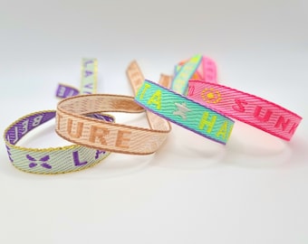 Op de afbeelding: Vijf kleurrijke geweven polsbandjes met verschillende tekst. De polsbandjes zijn roze, lichtblauw, beige en groen. De tekst op de polsbandjes is "Sunshine", "Hawaii", "Adventure", "Love" en "Island".