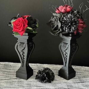 May include: Two black decorative pedestals with floral arrangements. One features red roses and black buds, the other has dark red and black flowers with black branches. A black flower sits in front.