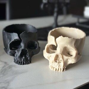 May include: Two skull-shaped planters. One is black, the other is off-white. Both have a cracked top edge and a hollow interior. The planters are on a white surface, with blurred decorative objects in the background.