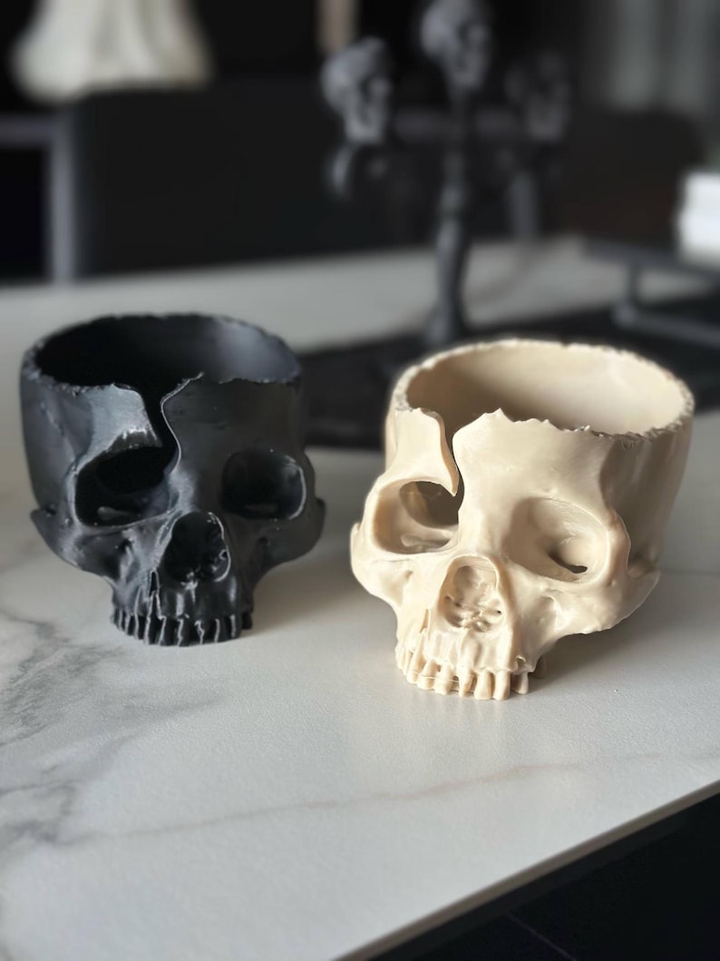May include: Two skull-shaped planters. One is black, the other is off-white. Both have a cracked top edge and a hollow interior. The planters are on a white surface, with blurred decorative objects in the background.