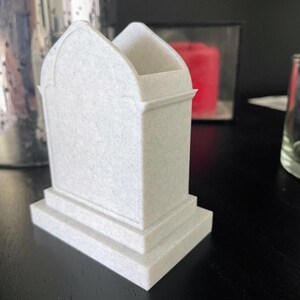 K&ouml;nnte beinhalten: Ein wei&szlig;er, kleiner Grabstein mit einer strukturierten Oberfl&auml;che und einem abgerundeten Bogen an der Oberseite. Der Grabstein hat einen Sockel mit zwei Stufen.