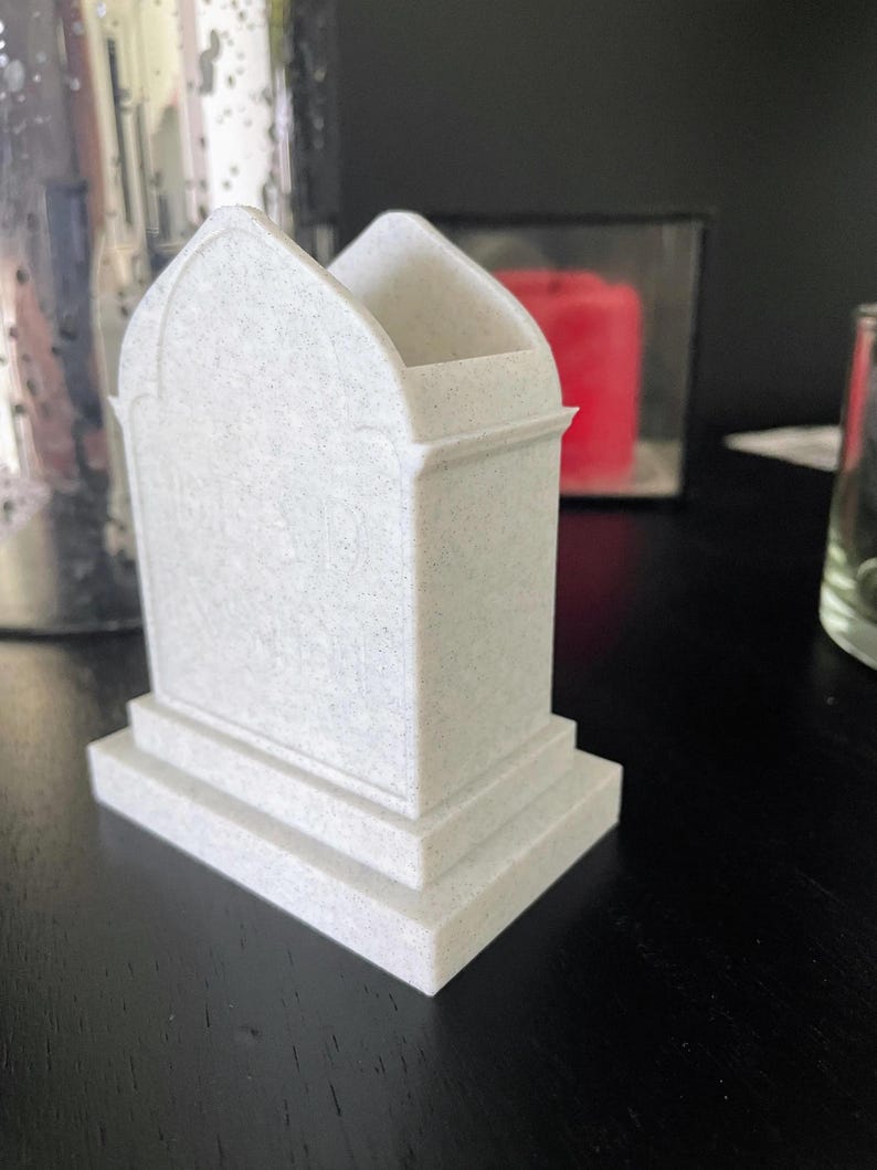 K&ouml;nnte beinhalten: Ein wei&szlig;er, kleiner Grabstein mit einer strukturierten Oberfl&auml;che und einem abgerundeten Bogen an der Oberseite. Der Grabstein hat einen Sockel mit zwei Stufen.