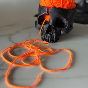 May include: A black skull ornament with a section of the top missing, filled with vibrant orange yarn. A length of the yarn trails from the eye. The skull sits on a light surface.