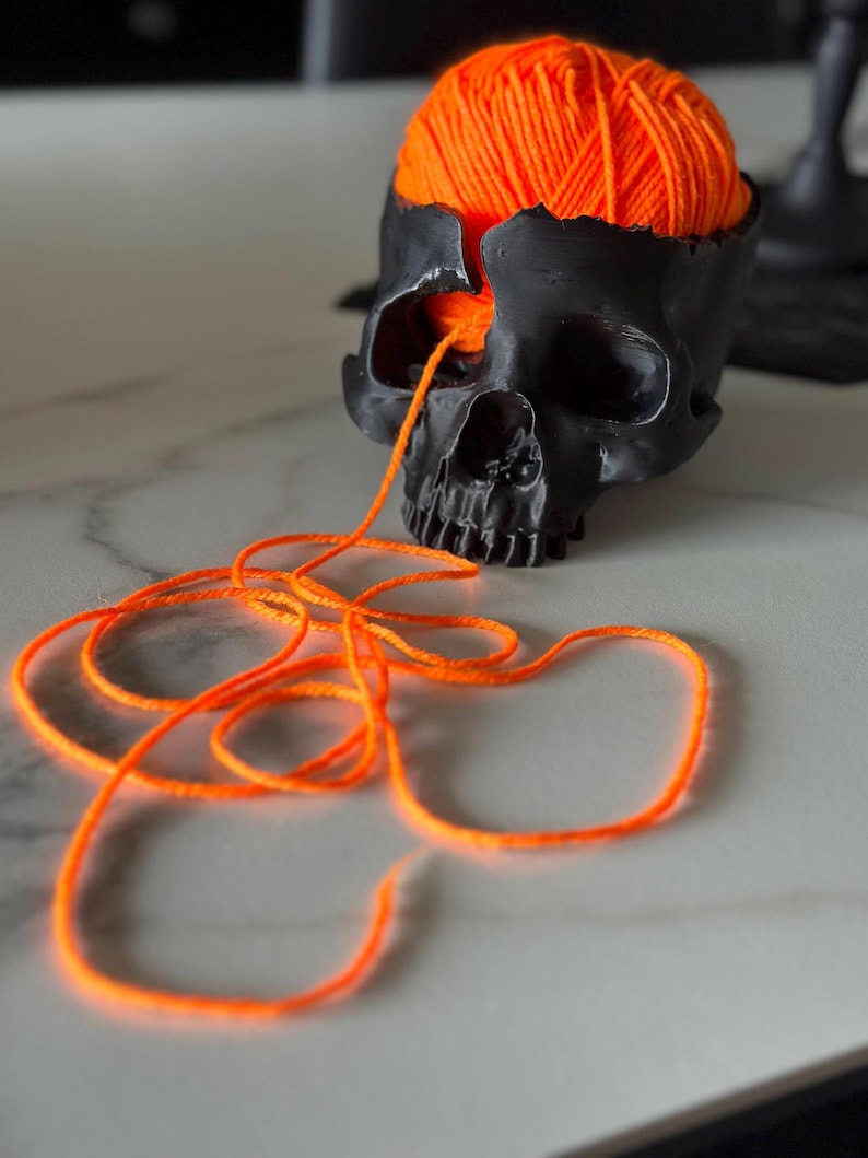 May include: A black skull-shaped container with a portion of the top removed, filled with bright orange yarn. A strand of the yarn extends from the eye socket. The skull is on a light-colored surface.
