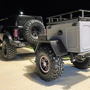 May include: Black Ford Bronco with a black and gray off-road trailer. The trailer has a roof rack and a gray box with two doors. The Bronco has a spare tire mounted on the back.