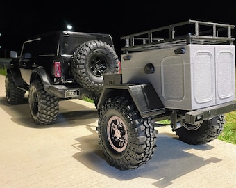 May include: Black Ford Bronco towing a black and grey off-road trailer. The trailer has a roof rack and a grey box with two doors. The Bronco has a spare tyre mounted on the back.