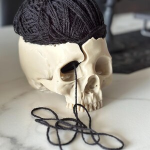 May include: A cream-colored skull with a dark black yarn ball resting on top. Black yarn threads extend from the skull's eye socket. The skull is on a white marble surface.
