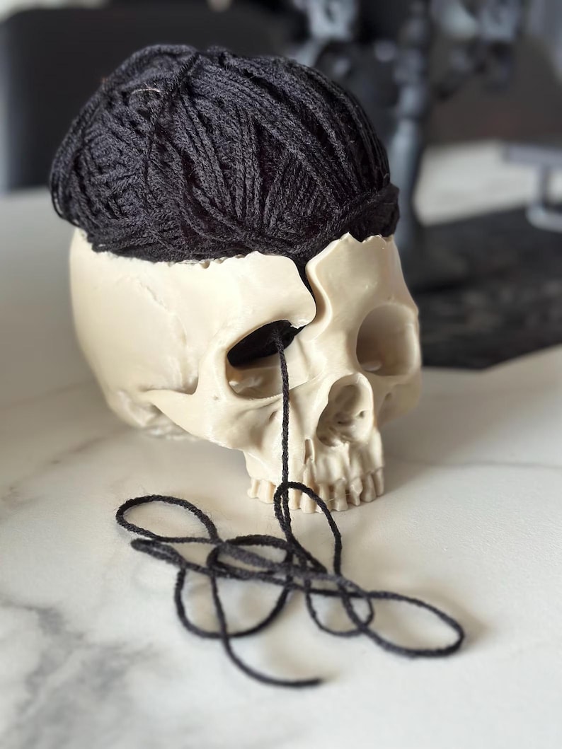 May include: A cream-colored skull with a dark black yarn ball resting on top. Black yarn threads extend from the skull's eye socket. The skull is on a white marble surface.