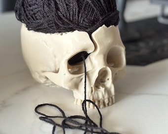 Peut inclure: Un crâne blanc avec une pelote de laine noire posée sur sa tête. La laine déborde des orbites et de la bouche du crâne.