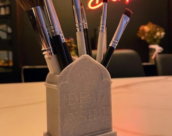 May include: A white ceramic makeup brush holder shaped like a tombstone with the words "Dead Inside" inscribed on it. Six makeup brushes are in the holder.