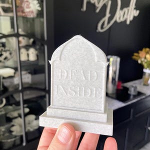 K&ouml;nnte beinhalten: Eine kleine, wei&szlig;e, sargf&ouml;rmige Dekoration mit der Aufschrift "DEAD INSIDE" auf der Vorderseite. Die Dekoration wird in der Hand einer Person gehalten.