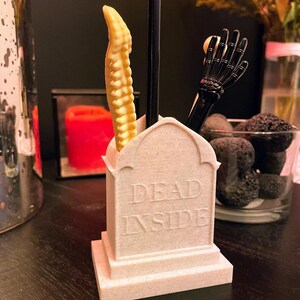 K&ouml;nnte beinhalten: Ein wei&szlig;er, keramik-tombsteinartiger Schreibtisch-Organizer mit den Worten "DEAD INSIDE" auf der Vorderseite. Der Organizer hat drei F&auml;cher f&uuml;r Stifte oder Bleistifte. Ein schwarzer, fledermausf&ouml;rmiger Stift, ein schwarzer, skelettartiger Handstift und ein gelber, skelettartiger R&uuml;ckenmarkstift aus Plastik befinden sich in den F&auml;chern.