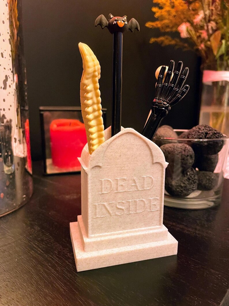 K&ouml;nnte beinhalten: Ein wei&szlig;er, keramik-tombsteinartiger Schreibtisch-Organizer mit den Worten "DEAD INSIDE" auf der Vorderseite. Der Organizer hat drei F&auml;cher f&uuml;r Stifte oder Bleistifte. Ein schwarzer, fledermausf&ouml;rmiger Stift, ein schwarzer, skelettartiger Handstift und ein gelber, skelettartiger R&uuml;ckenmarkstift aus Plastik befinden sich in den F&auml;chern.