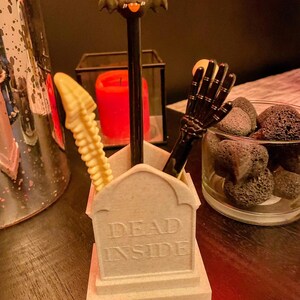 K&ouml;nnte beinhalten: Ein grauer, sargf&ouml;rmiger Schreibtisch-Organizer mit der Aufschrift "Dead Inside" auf der Vorderseite. Der Organizer enth&auml;lt einen schwarzen Stift mit einem Fledermaus-Topper, eine gelbe Skeletthand und eine schwarze Skeletthand.