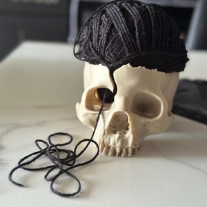 May include: A cream-colored skull with a black yarn ball on top, and a strand of yarn coming from the eye socket. The skull is on a white surface. The yarn is black.