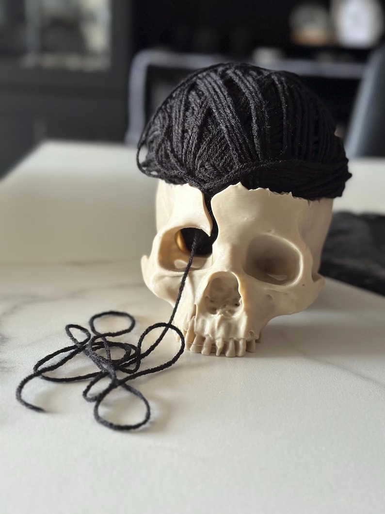May include: A cream-colored skull with a black yarn ball on top, and a strand of yarn coming from the eye socket. The skull is on a white surface. The yarn is black.