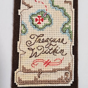 May include: A brown felt bookmark with a beige fabric map design. The map has a red cross, green and blue areas, and a red dashed line. The text "Treasure Within" is stitched in brown thread.