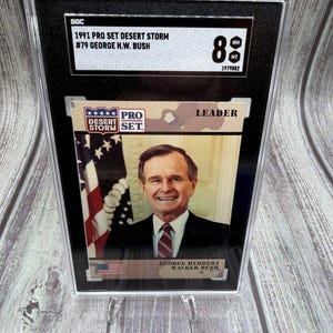 Könnte beinhalten: Eine bewertete Sammelkarte mit George H.W. Bush aus der 1991 Pro Set Desert Storm-Serie. Die Karte ist von SGC mit NM-MT 8 bewertet und enthält den Text "LEADER" und die amerikanische Flagge.