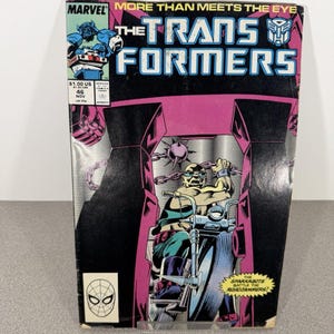 Puede incluir: Un cómic vintage titulado "The Transformers" con una portada vibrante. La portada presenta un personaje montando una motocicleta, sobre un fondo rosa y negro. El logotipo de Marvel y los detalles de la edición son visibles, junto con el texto "More Than Meets The Eye."