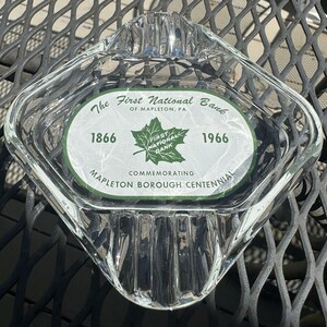 May include: A clear glass ashtray with a white oval label that reads "The First National Bank of Mapleton, PA. 1866 - 1966 Commemorating Mapleton Borough Centennial". The label features a green maple leaf with the words "First National Bank" inside.