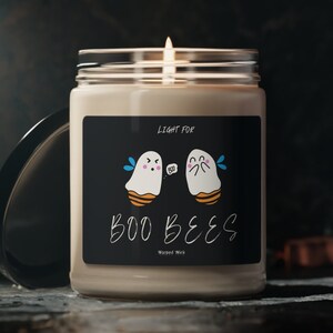 Pode incluir: Uma vela branca com um rótulo preto que diz "Light for Boo Bees" com um desenho de fantasma e abelha de desenho animado.