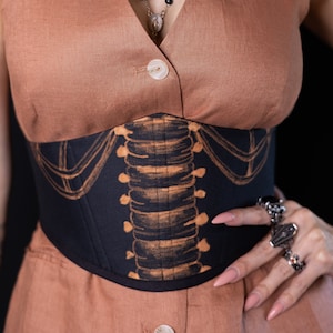 Könnte beinhalten: Ein schwarzes Korsett mit einem beigefarbenen Rippenmuster. Das Korsett wird über einem braunen Oberteil getragen. Das Model trägt Ringe und eine Halskette. Ein einzigartiges Modeaccessoire.