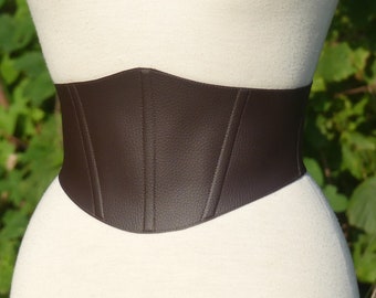 May include: A wide, dark brown corset belt featuring a textured, pebbled surface. The belt has a structured, contoured shape with vertical seam detailing across the front, designed to fit snugly around the waistline of a mannequin.