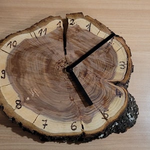 Könnte beinhalten: Eine rustikale Holzuhr aus einem Querschnitt eines Baumstamms. Das Zifferblatt hat schwarze Ziffern und Zeiger mit natürlicher Holzmaserung. Die Uhr hat einen Durchmesser von etwa 25 cm.
