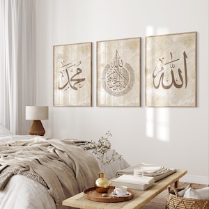 Puede incluir: Tres impresiones artísticas enmarcadas con caligrafía árabe en tonos marrones. Las impresiones se exhiben en una pared blanca sobre una cama con ropa de cama de color neutro. Un banco de madera con objetos decorativos está frente a la cama.