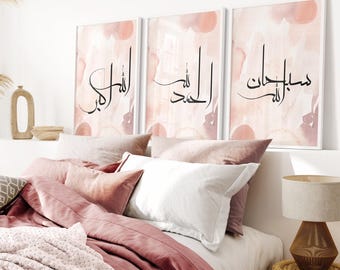 May include: Three framed art prints with Arabic calligraphy on a watercolor background in shades of pink and peach. The prints are displayed above a bed with pink bedding and pillows. A lamp with a woven base sits on a side table.