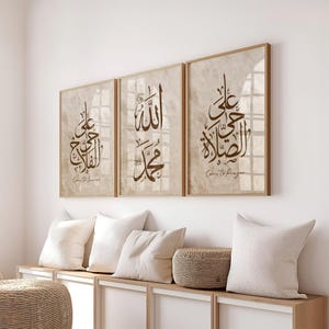 May include: Three framed art prints with brown calligraphy on a beige background. The prints are in wooden frames and feature Arabic script. The text "Come to Prayer" is at the bottom of two prints. The prints are displayed above white pillows and a woven basket.