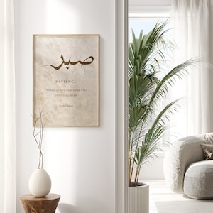 May include: A beige framed print with brown Arabic calligraphy that reads "Sabr" which means patience in English. The print also includes the text "Indeed Allah is with those who patiently endure" and "Quran 2:153".