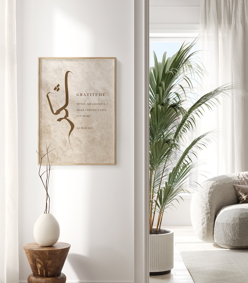 May include: A beige framed print with a brown calligraphy design and the text "Gratitude" and a quote from the Quran. The print is hanging on a white wall in a modern interior.