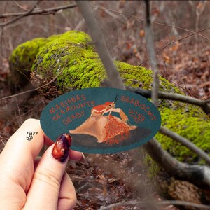 May include: An oval-shaped, teal-colored sticker with the text "MARIANAS SEA MOUNTS DERBY" and "SEABISCUIT WINS". The sticker features a crab illustration. The sticker is approximately 3 inches in diameter.