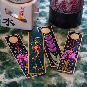 Könnte beinhalten: Vier Lesezeichen mit verschiedenen Designs. Ein Lesezeichen zeigt eine Spinne in einem Netz, ein anderes eine lila Blume, ein anderes eine rosa Blume und das letzte einen Mond und Sterne.
