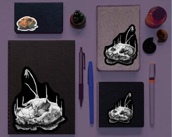 May include: A selection of notebooks and journals, sized from 7.6 to 22.9 cm, decorated with stickers. The stickers display a detailed black and white skull illustration. Also included are pens, a small potted plant, and decorative items.