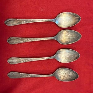 May include: Four antique silver spoons with floral detailing on the handles and oval bowls. The spoons are arranged in a row against a red background, showcasing their vintage design.