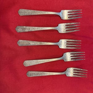 Puede incluir: Cinco tenedores plateados con mangos ornamentados sobre un fondo rojo. Cada tenedor tiene cuatro púas y un patrón decorativo en el mango. Los tenedores son de metal brillante, posiblemente cubertería antigua.