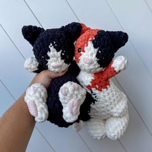 May include: Two handmade crochet cat plushies. One is black and white, the other is calico with black, white, and orange. Both have pink noses and paw pads. The plushies are soft and cuddly, ideal for a child's toy or a cat lover's gift.