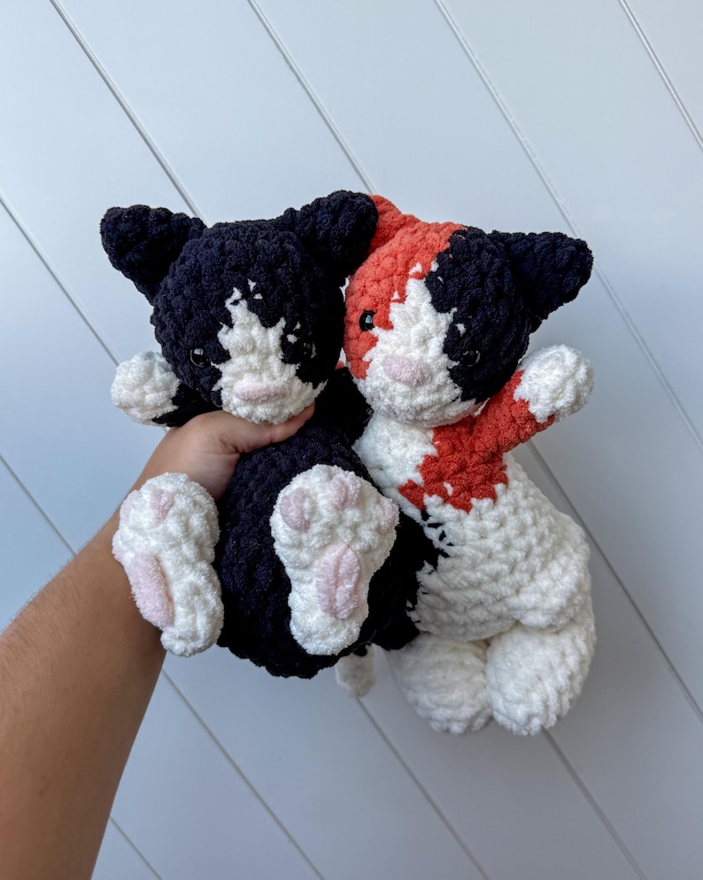 May include: Two handmade crochet cat plushies. One is black and white, the other is calico with black, white, and orange. Both have pink noses and paw pads. The plushies are soft and cuddly, ideal for a child's toy or a cat lover's gift.
