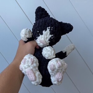 May include: A black and white crochet cat plush toy with pink paw pads. The cat has a white face and chest, with black ears and a black body. The plushie is soft and cuddly, ideal as a child's toy or a gift for a cat lover.