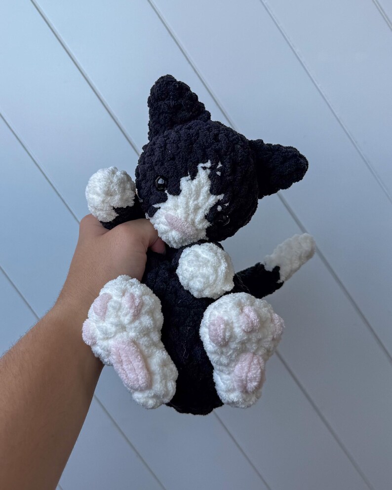 May include: A black and white crochet cat plush toy with pink paw pads. The cat has a white face and chest, with black ears and a black body. The plushie is soft and cuddly, ideal as a child's toy or a gift for a cat lover.