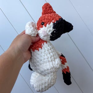 May include: A handmade crochet fox plush toy. The fox has a red head and tail, black ears and accents, and a white body with red and black markings. The fox has small black eyes and pink cheeks.