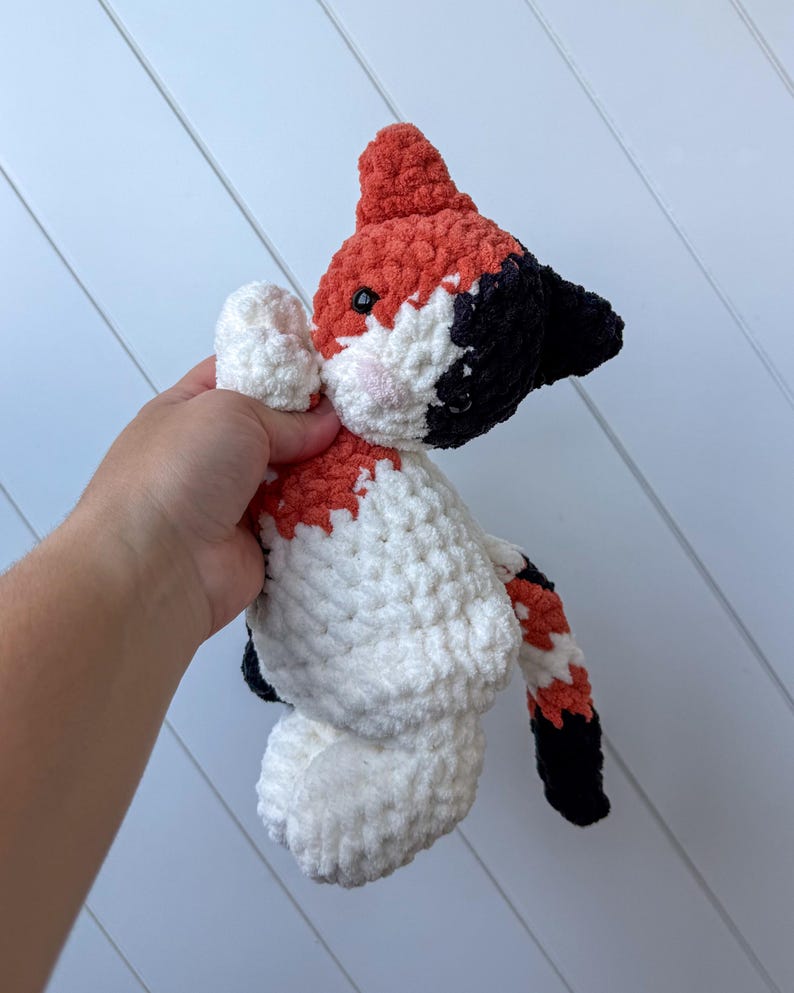 May include: A handmade crochet fox plush toy. The fox has a red head and tail, black ears and accents, and a white body with red and black markings. The fox has small black eyes and pink cheeks.