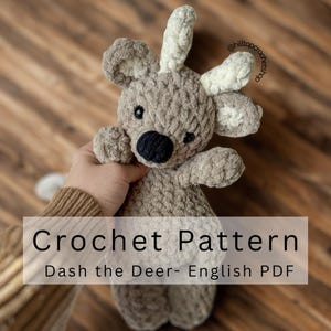 May include: A crocheted deer toy in grey and white. The deer has a dark grey nose, black eyes, and white antlers. The image includes the text "Crochet Pattern" and "Dash the Deer- English PDF".