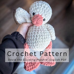 Peut inclure: Une peluche caneton au crochet faite à la main, en fil doux et texturé. Le caneton a un corps, des ailes et une tête gris clair, avec un bec et des pieds roses. Le texte "Crochet Pattern" et "Dovie the Duckling Plushie- English PDF" est affiché.